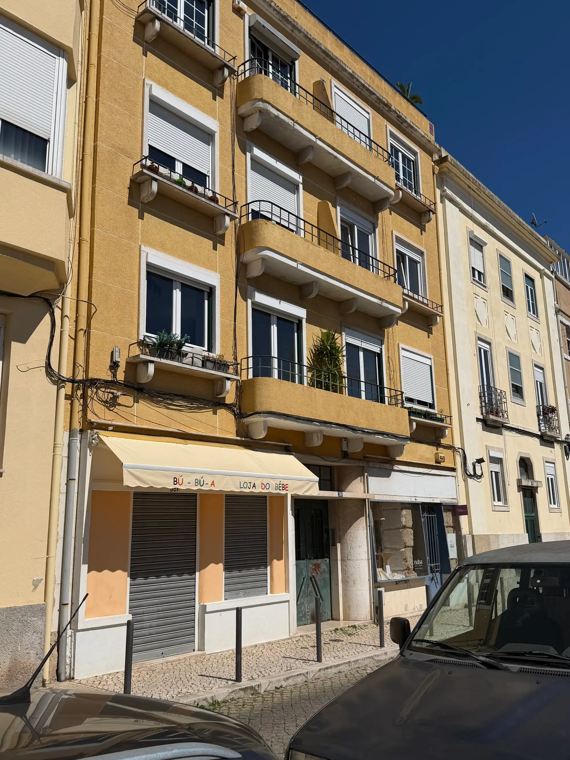 Fachada amarela de um prédio com varandas e uma loja fechada numa rua de calçada em Ajuda, Lisboa.