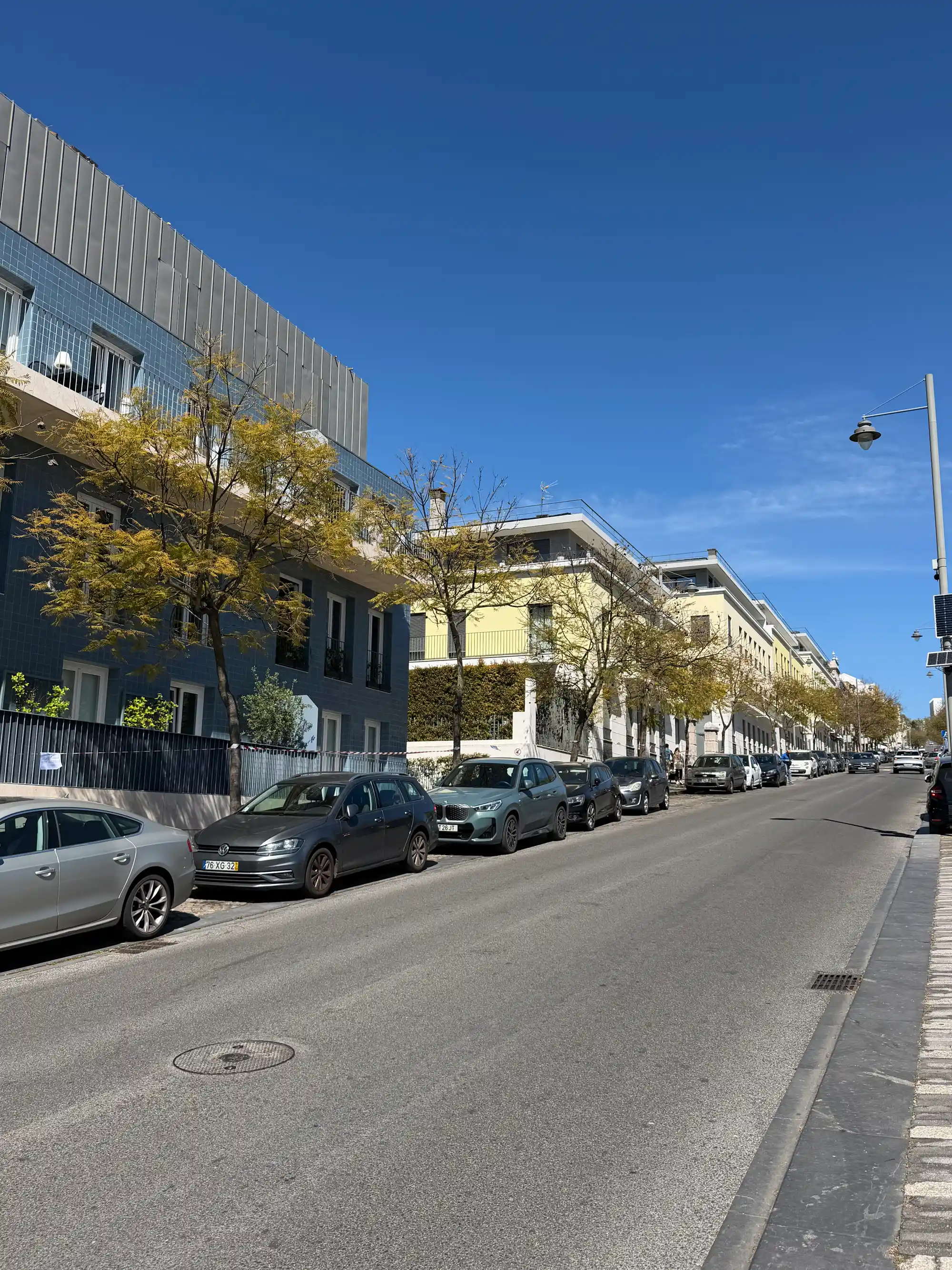 Rua em Ajuda, Lisboa, com carros estacionados, árvores e prédios residenciais com fachadas azuis e amarelas.