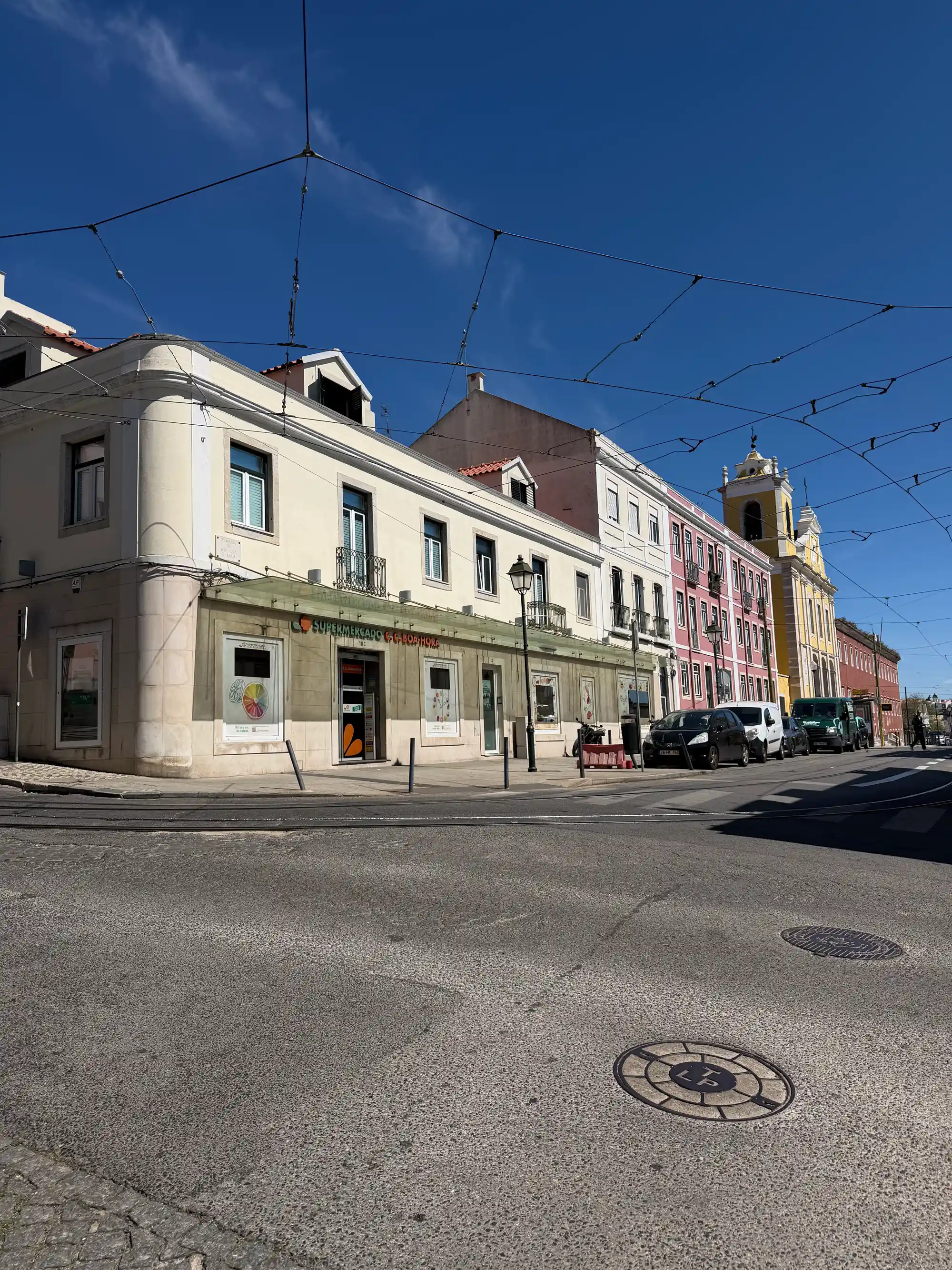 Esquina com supermercado e prédios coloridos perto da torre amarela de uma igreja em Ajuda, Lisboa.