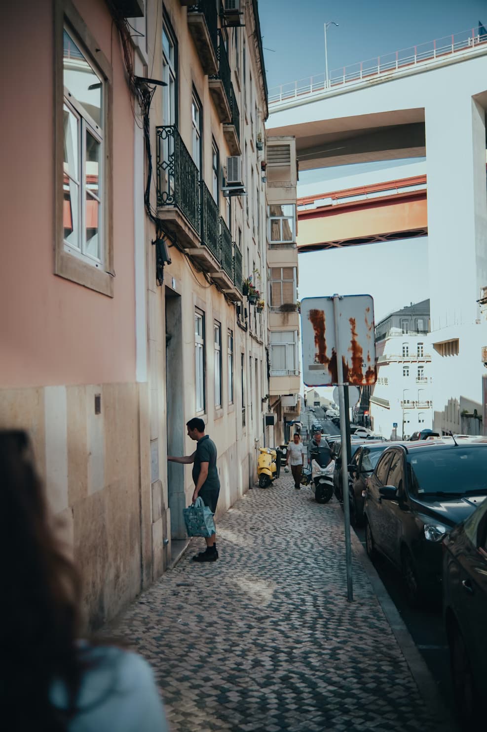 Alcantara: Homem entra em prédio por rua de paralelepípedos com ponte sobre ele.