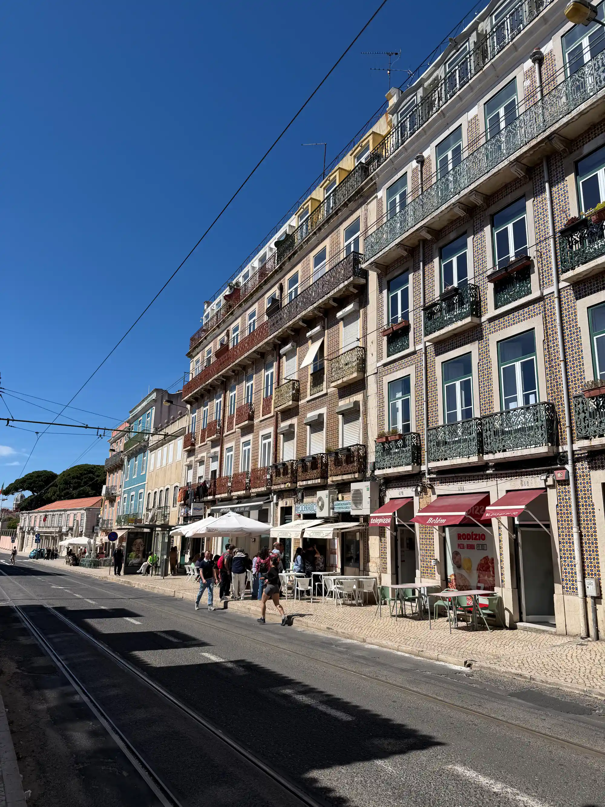 Vista da rua de edifícios revestidos de azulejos com varandas e mesas ao ar livre junto aos trilhos do elétrico em Belém, Lisboa.