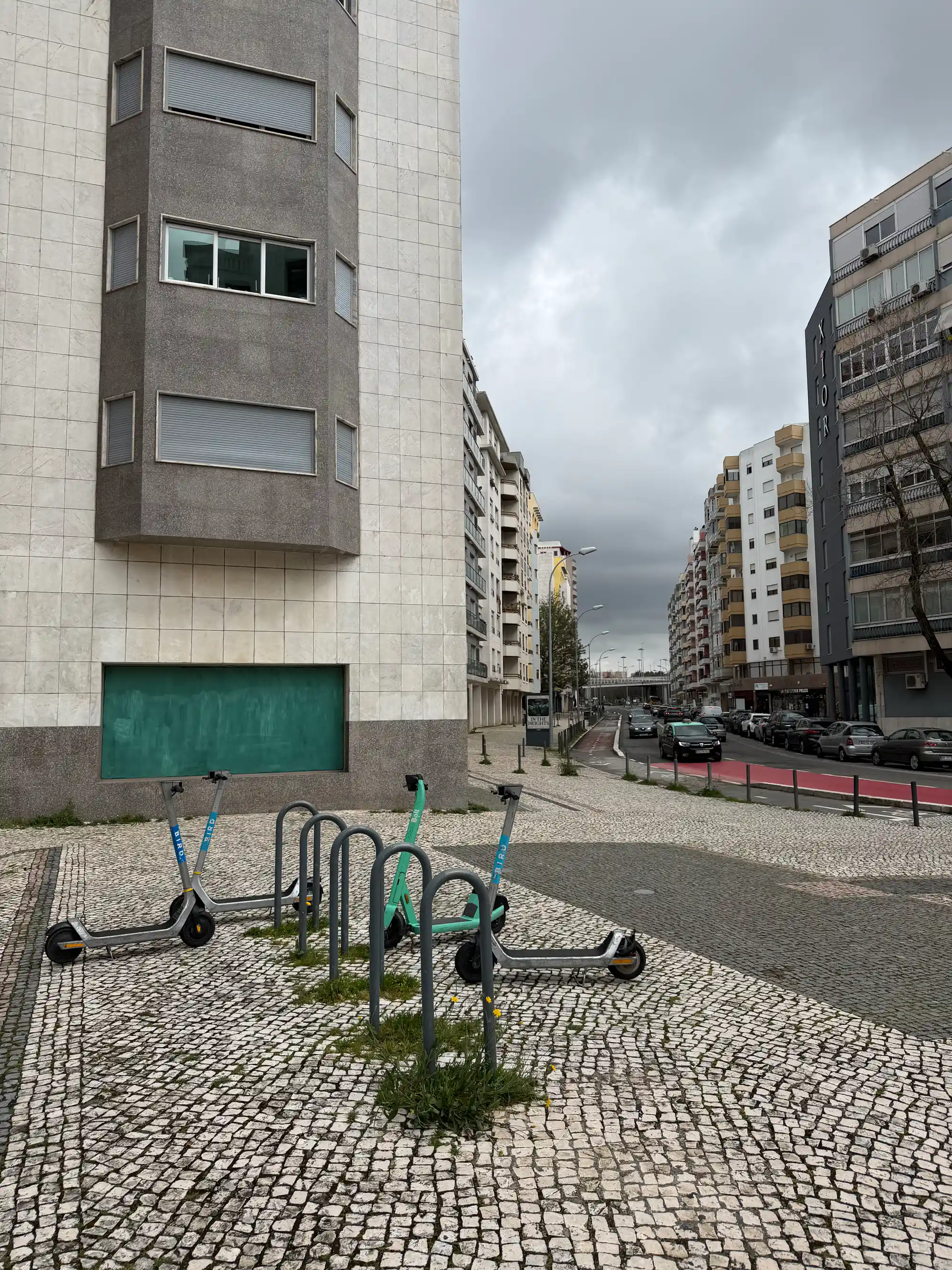 Rua residencial de Benfica, Lisboa, com prédios de apartamentos, carros estacionados e trotinetes elétricas partilhadas em ambiente urbano.