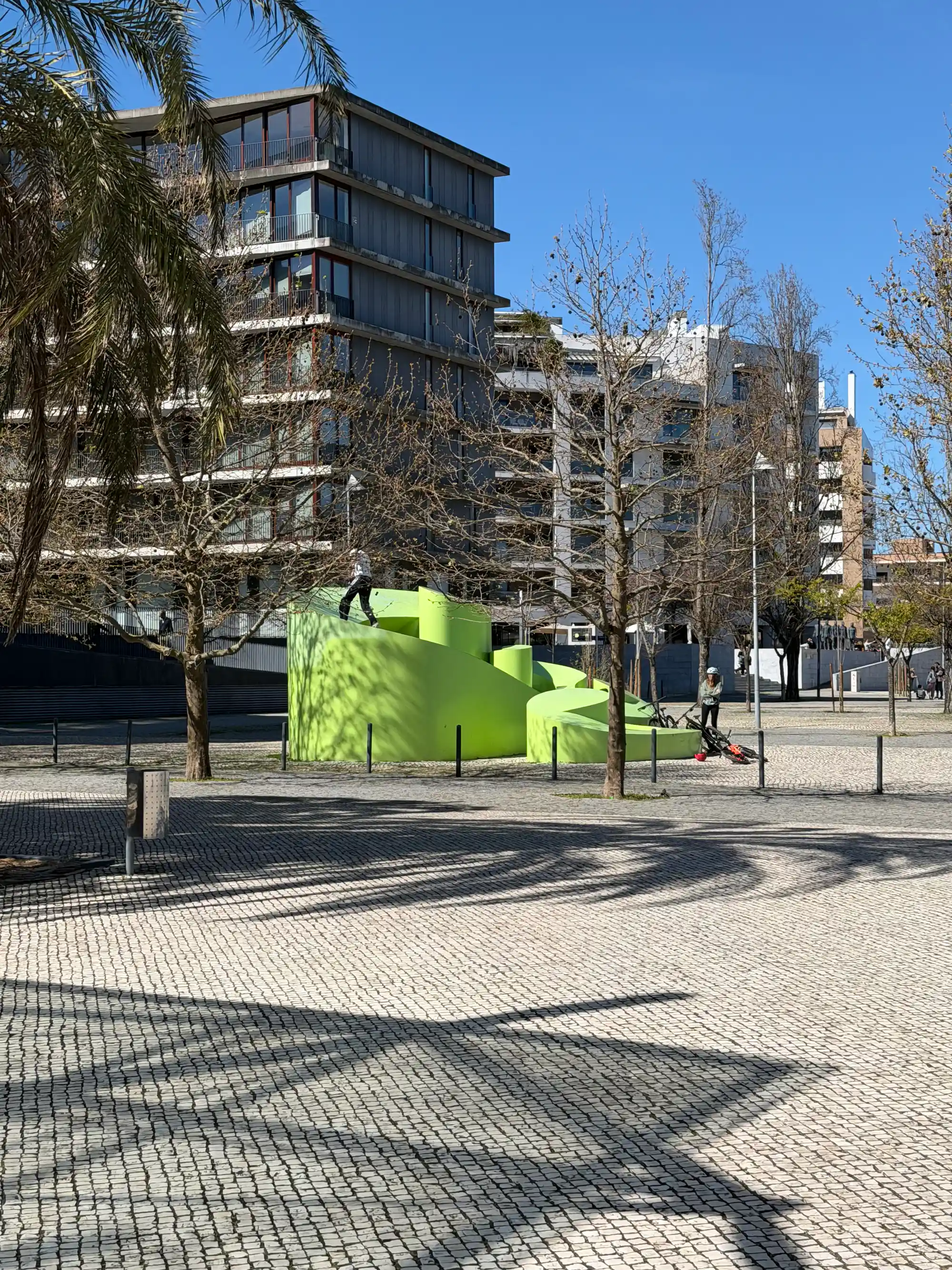 Escultura moderna em área verde no Parque das Nações, em Lisboa, com edifícios residenciais contemporâneos e sombras de palmeiras num dia ensolarado.