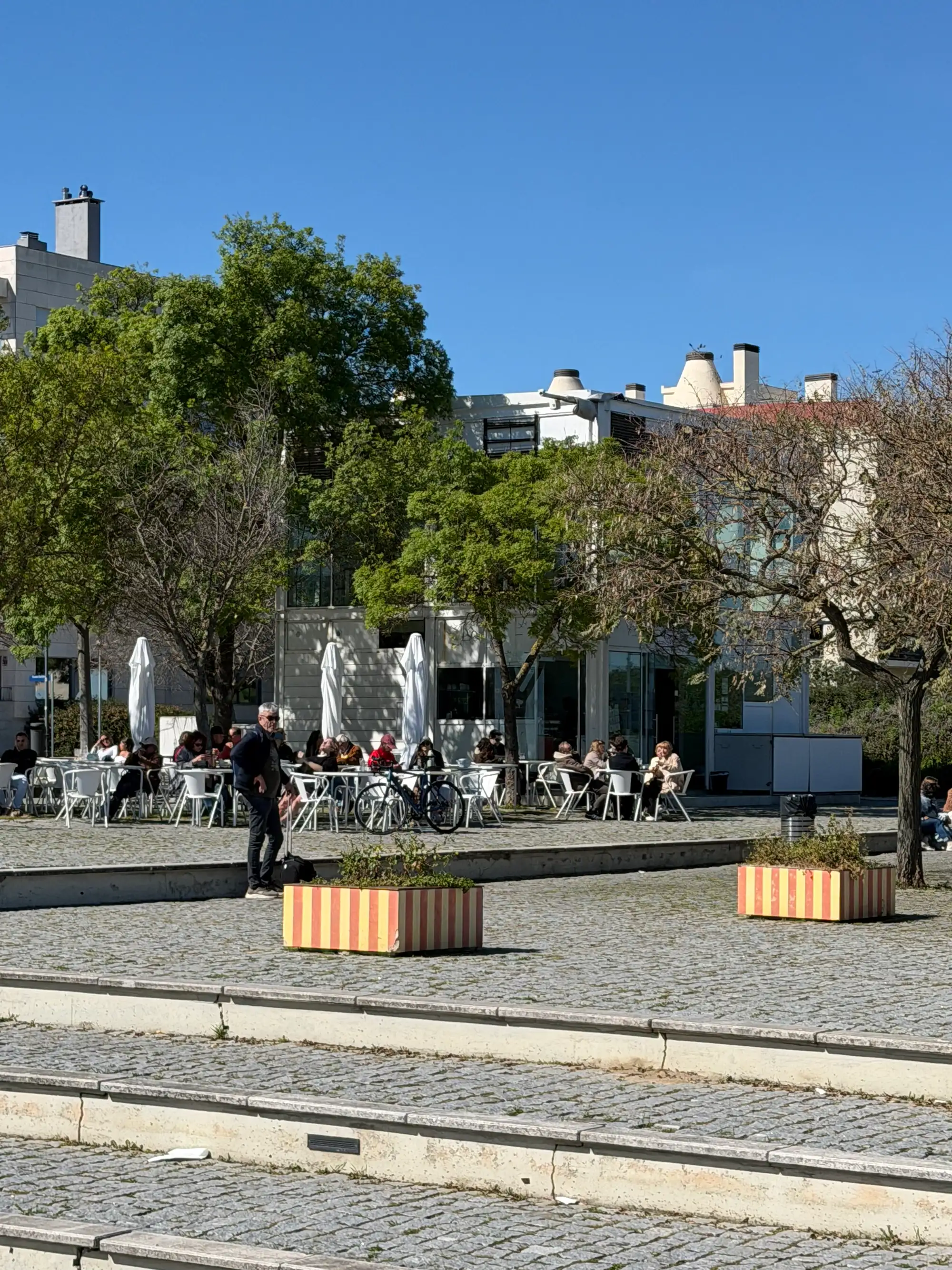 Terraço de um café ao ar livre com pessoas sentadas às mesas no Parque das Nações, em Lisboa, rodeado por árvores e edifícios modernos num dia ensolarado.